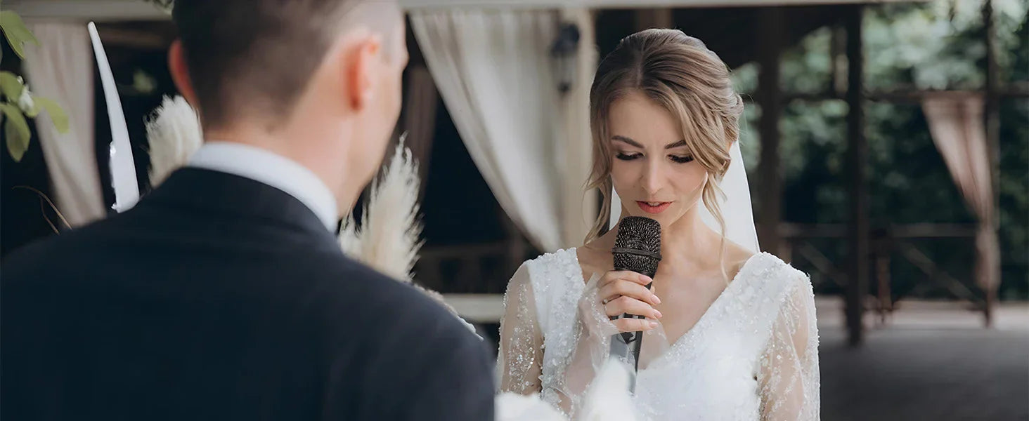 A bride in a white wedding gown speaks into a Talomen K60-2 dual wireless handheld dynamic microphone during a wedding ceremony, while the groom stands facing her.